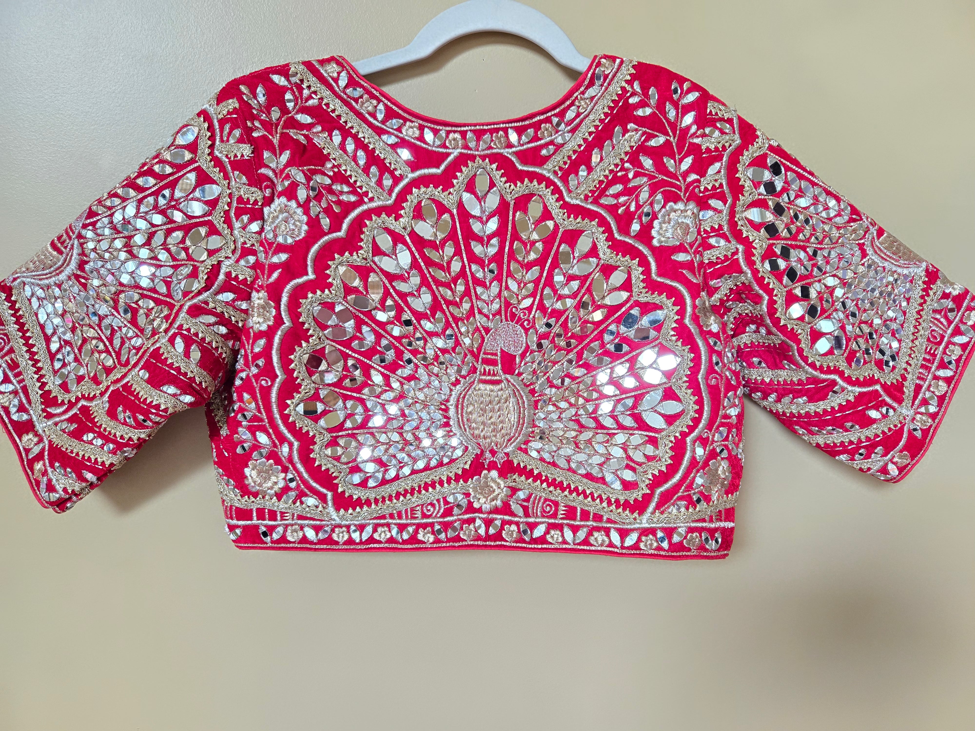 A bright pink embellished blouse top with extensive silver mirror work and embroidery, hanging on a white plastic hanger against a plain beige wall.
