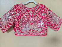 A bright pink embellished blouse top with extensive silver mirror work and embroidery, hanging on a white plastic hanger against a plain beige wall.